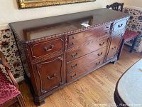 Front angle showing entire wooden dresser with ornate carved accents and metal drawer handles. Glossy top surface with scratches visible.