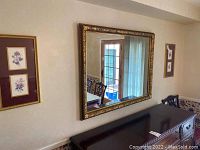 Wall mirror with golden finished frame hanging above a dark wooden sideboard, reflecting a window and chair.