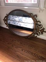 Oval mirror with detailed gold painted frame resting on hardwood floor beneath a window. Clearly shows the ornate curls and leaf motifs on the frame sides and the reflective surface.