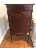 Closed view of vintage music cabinet showing the decorated front door with veneer missing on bottom right and a small drawer above.