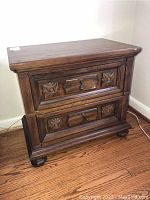 Front view of wood nightstand showing two drawers with carved floral designs and metal handles, sitting on short curved feet.