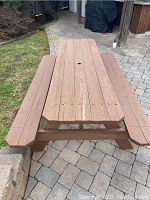 Front view of rectangular wooden picnic table with attached benches on a paved patio, showing weathered wood with surface wear and central umbrella hole.