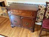 Front angled view of the wooden server showing two drawers, two cabinet doors, decorative carved details, and block feet on hardwood floor.