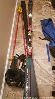 Photo shows two black Olin Radius skis about 78 inches long with attached bindings, two bright red Tomic T-4 ski poles with yellow grips leaning against the wall, black Nordica ski boots, and a blue ski boot bag on the floor.
