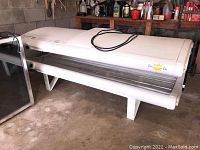 Full side view of white Sunmaster tanning bed with power cord on top, inside tanning tubes visible under hinged lid.