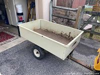 Full side view of cream-colored rectangular metal tractor cart showing rust and weathering. Mounted on wheels with hitch for vehicle attachment.