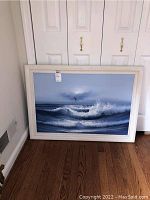 Framed ocean wave and seagull painting on floor leaning against wall, showing full artwork and frame with a label.