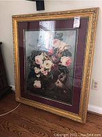 Framed floral print featuring painted flowers in a vase. The frame is gold-painted wood with decorative molding and covered with glass. Print is matted in burgundy and beige. Visible in a well-lit interior setting.