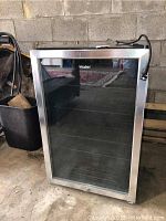 Front view of the Haier beverage center with glass door and metal frame. Unit is standing on a concrete floor showing signs of rust and stains.