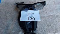 Black covert video recording glasses positioned on top of a black zippered protective case, both photographed against a light surface with a lot label number in the center.