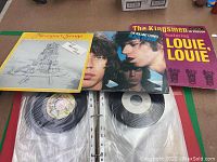 Photo of three vinyl albums on top of binder containing 45 RPM records, showing The Kingsmen, The Rolling Stones, and Cheryl Wheeler albums.