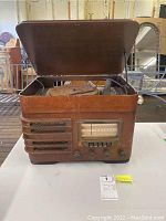 Front view of the wooden cabinet with open hinged lid, showing radio tuner with dial and control knobs, and horizontal vent openings on front left side.