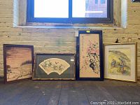 All four framed Asian-style artworks positioned on the floor against a wall under a window.