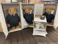Three teddy bears dressed in black suits, seated inside original boxes on wood surface. The bears have light brown fur and are all 16 inches tall. To the right is a smaller white poodle doll in its box.