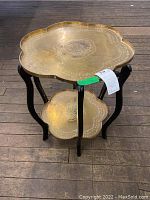Full view of brass folding tray table on wooden floor, showing two scalloped brass trays and black wooden legs.