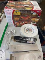 Photo showing Kitchen Delight 15-piece glass ovenware boxed set, stack of nine white Wedgwood plates with floral central pattern, two boxed silver plated salad serving sets and silver plated tongs.