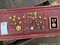 Top-down photo showing the full assortment of military medals, buttons, pins, and a coin spread on a red surface. Includes gold-colored buttons, enamel pins, ribbons, and a boxed medal.