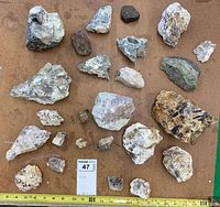 Top-down view showing 25 different rocks and mineral specimens arranged on a brown surface with a yellow measuring tape below for scale.