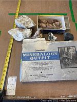 Full view of box with original label, several mineral specimens outdoors from tray, several loose rocks beside box