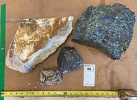 Four assorted rocks and minerals arranged on a brown surface with measuring tape showing size up to about 12 inches, including large beige rock, large dark bluish-black rock, small dark rock, and mid-sized speckled rock.