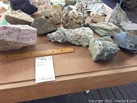 Photo showing a variety of over 25 different rocks and mineral specimens laid out on a table, with a wooden ruler for scale.