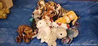 Photo showing multiple stuffed animals grouped on a blue tarp including several tiger TY Beanie Babies, two Hello Kitty plush toys with red bows, a white bunny, a white bear, an elephant, and a dog plush.