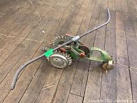 Wide angle photo showing full walking sprinkler with metal pipe arms, toothed gear wheel, and front wheel on wooden floor.