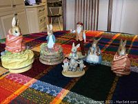 Full lot of six Beatrix Potter themed figurines on a plaid cloth background showing various characters including rabbits in cloaks and a human figure.