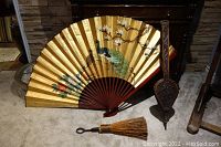 Photo showing large decorative fan with peacock and floral artwork, metal bellows, and small whisk brush arranged on carpeted floor.