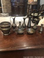 Front view of seven brass decorative pieces on wooden table including various ewers, bowls, and an incense burner.