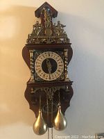 Front view of wooden and brass wall clock showing clock face, weights, and decorations.