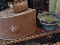 Photo of copper corner pedestal, planter, and decorative metal bowl on a carved wooden surface.
