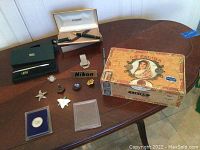 Overview of lot: two boxed pens, metal pencil, La Palina cigar box, Nikon badge, coin, money clip and assorted small decorative items