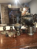 Photo showing entire lot on wooden table with background furniture, displaying all brass items including vases, candle holders, mug, and figurines.