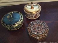 Three decorative containers displayed closed on a wooden surface: blue glass and brass jar, ceramic jar with gold lid and classical figures, octagonal wooden inlaid box.
