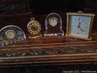 Four small desk clocks displayed on a wooden surface. Includes varied designs in metal and glass with wear visible.
