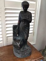 Front and side views of the standing female fountain statue showing the figure, water basin with lion's head spout, and textured base on a wooden surface near a window.