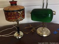Two table lamps side by side on a wood table; left lamp with Kodak stained glass shade, right banker lamp with green glass shade