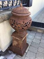 Full view of ornate metal garden urn with pedestal and lid on stone pavers