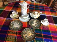 Photo showing various collector plates, teacups, teapot, and a tall covered pitcher arranged on a colorful plaid tablecloth.