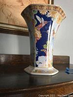 View of hexagonal vase showing blue panel with bird and flower design, gold trim on rim.
