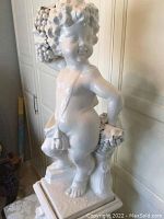 Front-left side view of white ceramic cherub statue holding grapes, standing on ornate pedestal base.