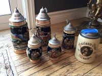 Six Western Germany beer steins displayed on wooden and textile surfaces, showing size range and painted scenes.
