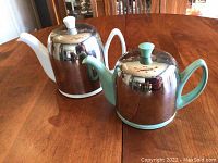 Two vintage tea and coffee pots side by side on wooden table, showing metal bodies with different colored handles and lids.