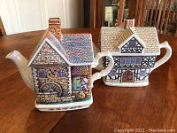Two ceramic teapots designed as detailed houses sitting on a wooden table, showing side angles and articulations of spouts and handles.