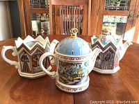 Three Sadler teapots displayed on wood table: one blue domed fishing scene teapot and two crown-shaped portrait teapots
