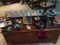 Frontal view of multiple vintage cameras on wooden chest with light blue 'CAMERAS' sign