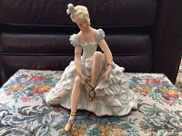 Full frontal view of the seated ballerina figurine showing her adjusting her shoe, dressed in ruffled pale blue and white dress with gold ballet slippers.