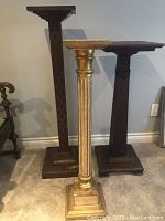 Photo of three wood display stands including the gilded fluted column and two carved dark wood stands, showing overall condition with scratches and wear.