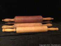 Four antique wooden rolling pins displayed horizontally on a black surface showing varied wood colors and handles